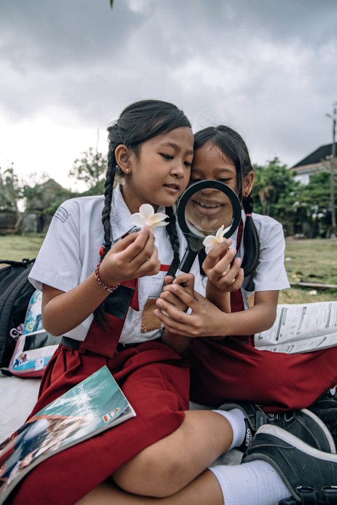 Two schoolchildren in uniforms observing flowers with a magnifying glass outdoors.