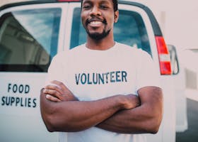 African American volunteer smiling in front of food supplies van with arms crossed, promoting community service.